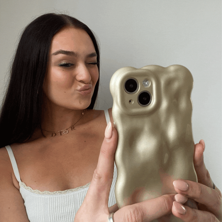 A woman with long dark hair, wearing a white tank top and a "Dior" necklace, is taking a selfie with her iPhone15 adorned in Coconut Lane's 3D Pearl Phone Case - Gold. She is winking and making a playful duck face expression near a white background.