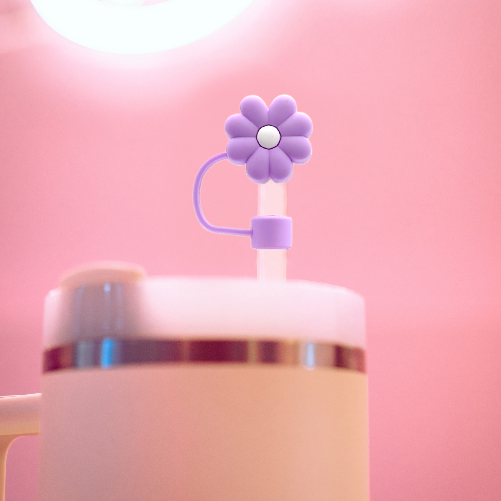 A cream-colored tumbler with a metallic band and a lid featuring a clear straw, accentuated by Coconut Lane's Lilac Flower Straw Cover. The background is softly lit with a warm pink hue.
