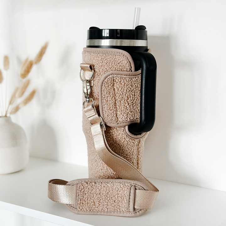 The Teddy Rupert Tumbler Carry Case by Coconut Lane, featuring a beige textured design and a strap, is displayed on a white surface. Accompanying it is a large black-handled travel mug with a straw emerging from its lid. In the background, there's a white vase containing dried wheat stalks.