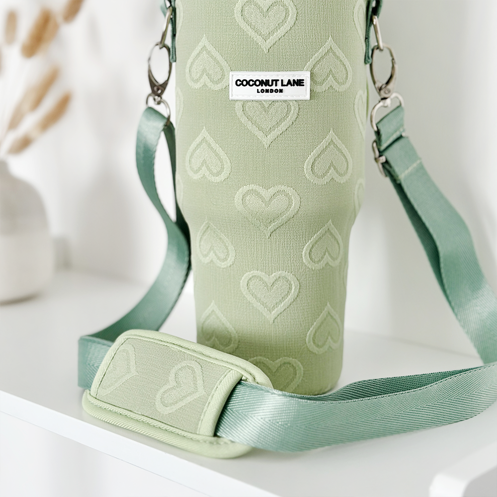 A Tumbler Carry Case - Sage Heart by Coconut Lane, characterized by its light green hue and heart pattern, is showcased on a white surface. The bag features a detachable strap and bears a small rectangular label reading "Coconut Lane London." It is positioned in front of a white vase with dried flowers, which appear slightly out of focus in the background.