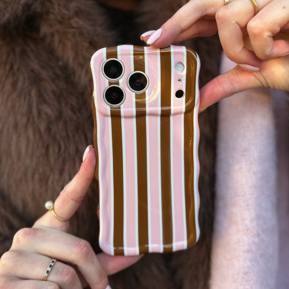 Woman holding an iphone in a cocoa & Pink Stripe Wavy Phone Case featuring glossy brown, blush pink and cream vertical stripes, photographed diagonally on a pink background. She is wearing a baby pink scarf and faux fur brown jacket.