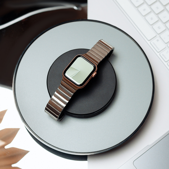 A smartwatch with a Coconut Lane Slinky Apple Watch Strap - Silver rests on a circular wireless charging pad. The charger sits atop a silver laptop, and part of a geometric patterned surface can be seen in the background.