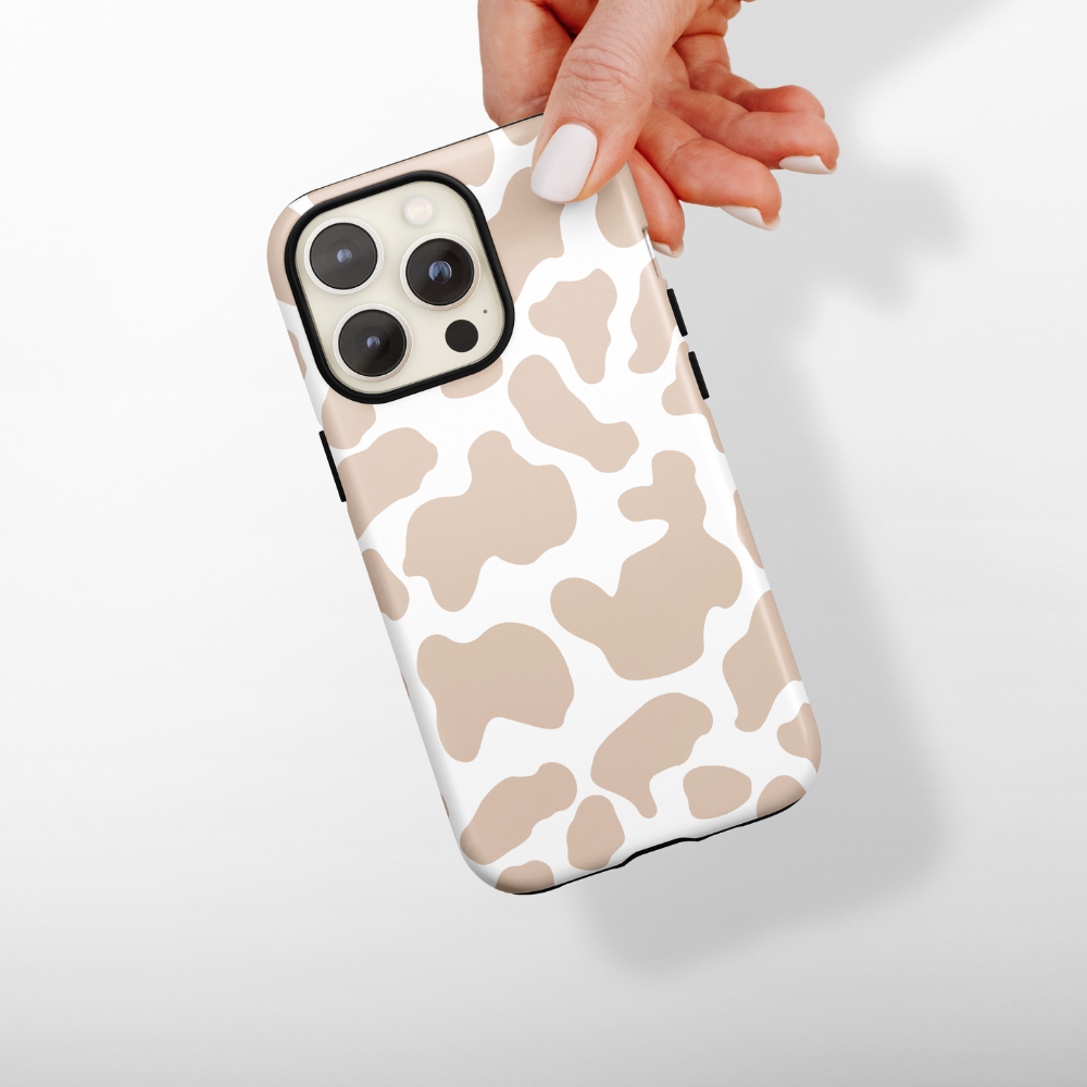 A hand with a white manicured thumb holds an iPhone encased in the Coconut Lane "Tough Phone Case - Nude Cow." The background is plain white, casting a soft shadow behind the phone.