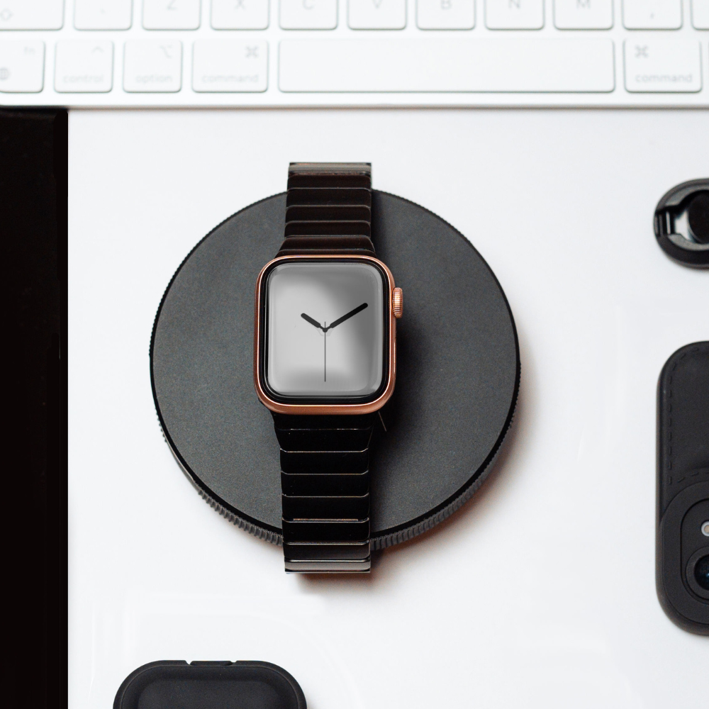 A smartwatch with a Coconut Lane Slinky Apple Watch Strap - Black is placed on a black wireless charging pad. Behind the watch, a section of a white keyboard is visible, and to the right, there are a camera lens cap and part of an electronic device. The background surface is white.