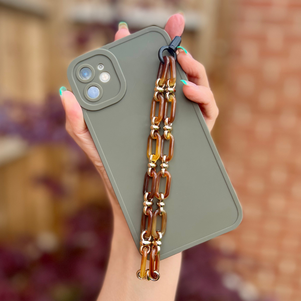 A person's hand is holding a green smartphone encased in a minimalist Coconut Lane NAKD Phone Case - Khaki, offering full camera protection. Attached to the case is a decorative chain featuring interlinked brown and gold-tinted links. The background is blurred, showcasing wooden and brick textures, while colorful, manicured nails add a touch of flair.