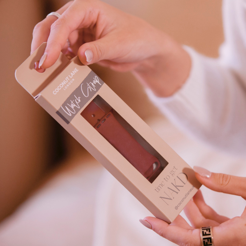 A person with long nails is holding packaging for a pink phone case. The packaging, labeled "Coconut Lane," "NAKD Apple Watch Strap - Red Wine," and "Time to Get Naked," reveals a sleek Silicone Strap designed for Apple Watch models as the hand slightly lifts the tab to open it.