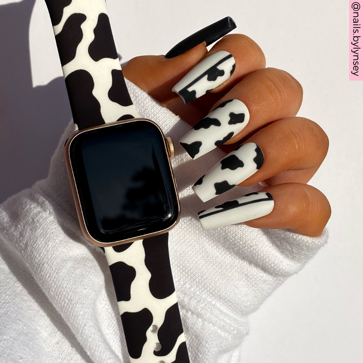 A person with a Coconut Lane Cow Apple Watch strap and a cow print manicure holds their smartwatch. The nails and watch band both feature a white background adorned with black cow-like spots. The person is wearing a white long-sleeve shirt, and the Instagram handle "@nailsbylynsey" is visible in the top right.