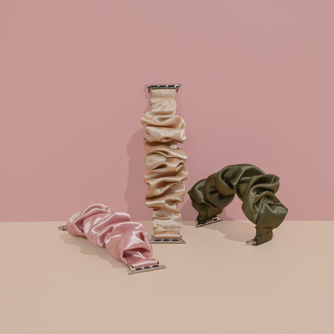Three scrunchie wrist bands for Apple Watch are showcased against a pink and beige backdrop. The silky satin bands, including the Vintage Khaki Scrunchie Apple Watch Strap from Coconut Lane along with soft pink and champagne gold variants, are arranged in a standing position, leaning against each other.