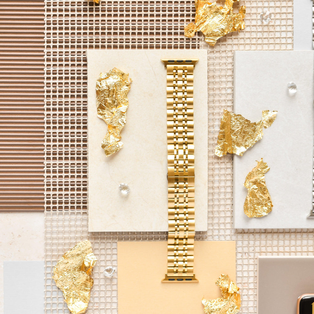 An overhead view of the Coconut Lane Stainless Steel Apple Watch Strap - Gold, elegantly placed on a combination of grid and solid backgrounds in beige and light brown tones. Gold leaf pieces and small clear beads are artfully scattered around the band.