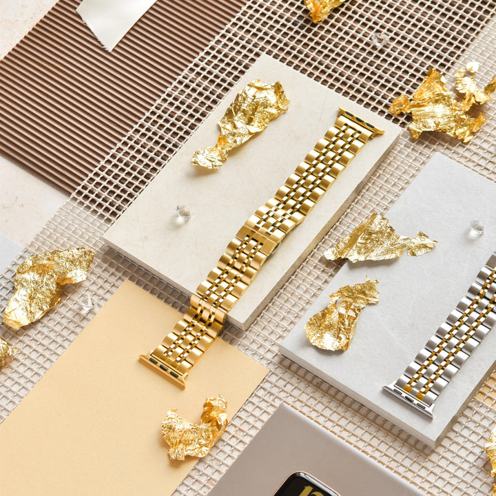 A decorative display featuring a Stainless Steel Apple Watch Strap - Gold by Coconut Lane and a silver adjustable Apple Watch band, arranged on textured fabric and paper backgrounds. The arrangement is adorned with small pieces of gold leaf, creating an elegant and artistic presentation.