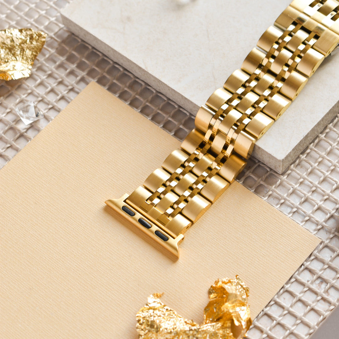 A Stainless Steel Apple Watch Strap - Gold by Coconut Lane is displayed on a beige and white tile background, atop a mesh surface. Small pieces of gold leaf are scattered around, adding a touch of elegance and luxury to the scene.