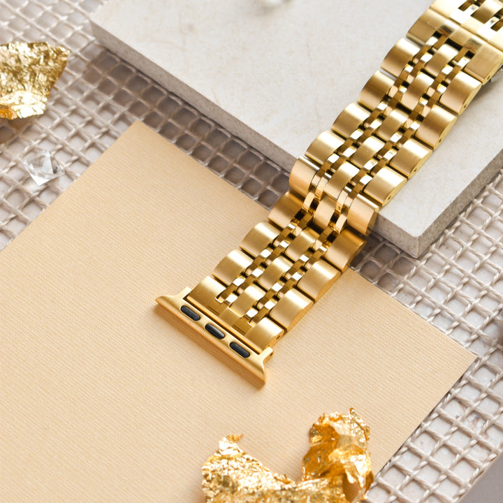 A Stainless Steel Apple Watch Strap - Gold by Coconut Lane is displayed on a beige and white tile background, atop a mesh surface. Small pieces of gold leaf are scattered around, adding a touch of elegance and luxury to the scene.