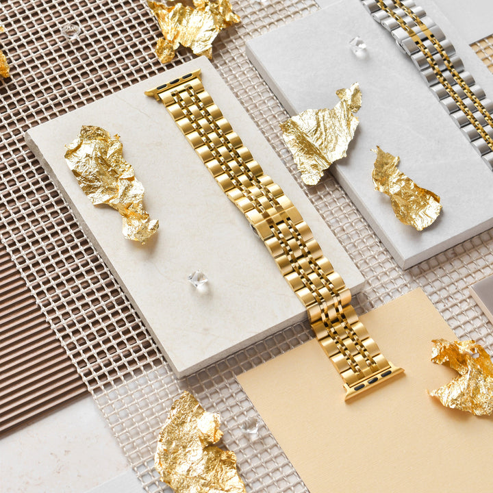 A Stainless Steel Apple Watch Strap - Gold from Coconut Lane lies horizontally on a geometric display of various textured panels including beige, white, and mesh materials. Gold foil pieces are scattered around, enhancing the luxurious aesthetic of the setup.