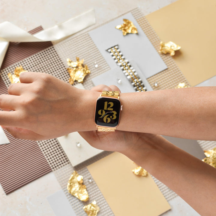 A person wearing a gold smartwatch with a black face displaying the time "12:36". The background features various textured materials in neutral colors, along with gold foil accents scattered around. The person’s other hand is adjusting the Coconut Lane Stainless Steel Apple Watch Strap - Gold on their wrist.