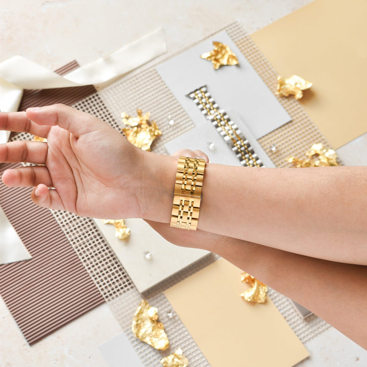 A hand adorned with Coconut Lane’s stylish Stainless Steel Apple Watch Strap in Gold is showcased against a backdrop of various materials and gold foil accents. The scene features textured paper, ribbons, and decorative beads, creating a sophisticated and elegant setting.
