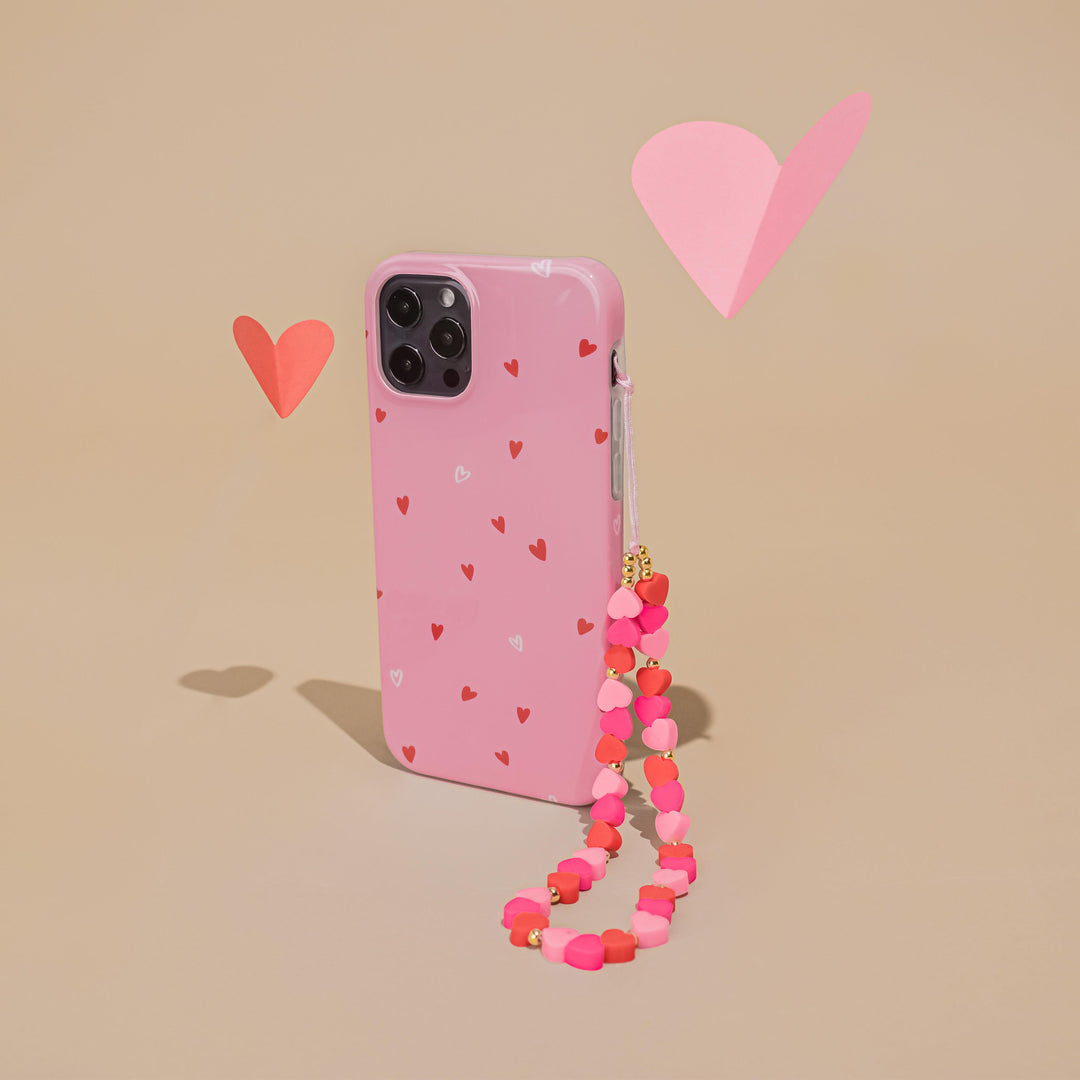 A pink phone case adorned with small red and white hearts stands upright against a beige background. Attached to the case is Coconut Lane's Beaded Phone Strap - Hearts in pink and red. Two heart-shaped paper cutouts in red and pink float nearby.