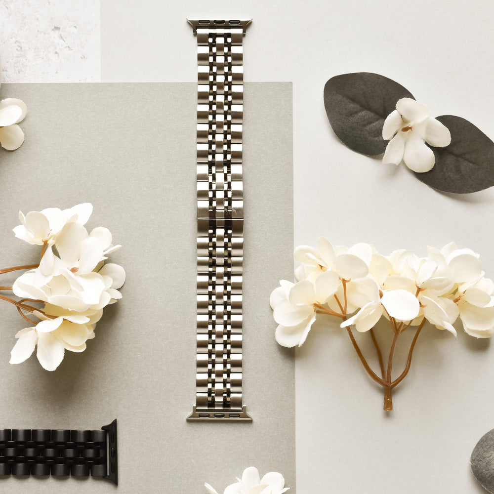 A Coconut Lane Stainless Steel Apple Watch Strap - Black & Silver is displayed in the center of the image on a light gray background. Surrounding it are white flowers and a dark green pair of leaves. Another black watch band, compatible with various Apple Watch models, is partially visible at the bottom left corner.