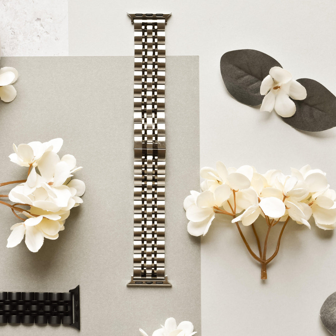 A Coconut Lane Stainless Steel Apple Watch Strap - Black & Silver is displayed in the center of the image on a light gray background. Surrounding it are white flowers and a dark green pair of leaves. Another black watch band, compatible with various Apple Watch models, is partially visible at the bottom left corner.