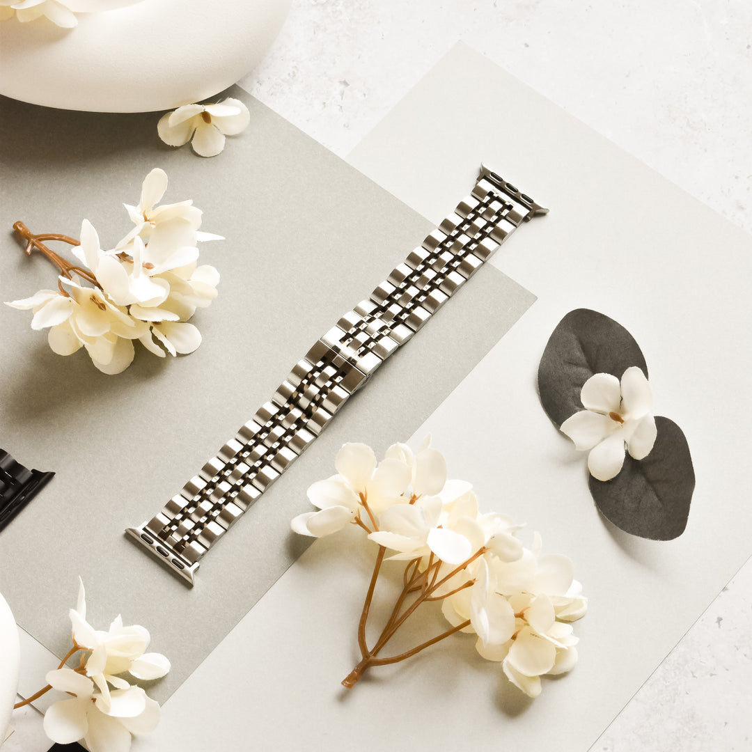 A Stainless Steel Apple Watch Strap - Black & Silver from Coconut Lane is placed on a light gray and white surface, surrounded by white artificial flowers. Two black and gray watchbands are partially visible on the sides, highlighting the adjustable metal band in the center, compatible with various Apple Watch models.