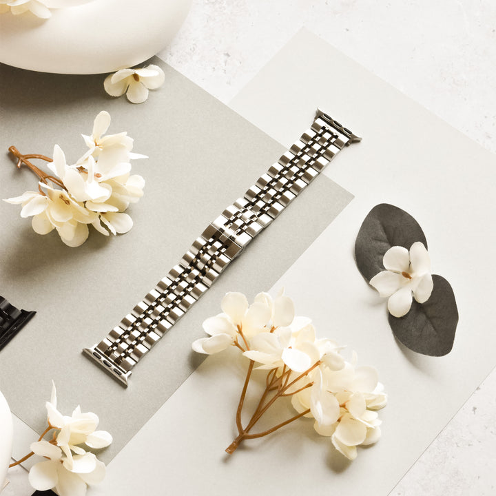 A Stainless Steel Apple Watch Strap - Black & Silver from Coconut Lane is placed on a light gray and white surface, surrounded by white artificial flowers. Two black and gray watchbands are partially visible on the sides, highlighting the adjustable metal band in the center, compatible with various Apple Watch models.