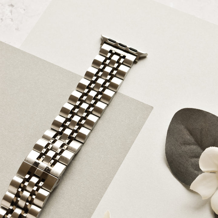 The Coconut Lane Stainless Steel Apple Watch Strap - Black & Silver, featuring a sleek, segmented design, is laid out on a light-colored surface. Nearby, a dark gray leaf and a small white flower add a touch of natural contrast to the modern accessory.