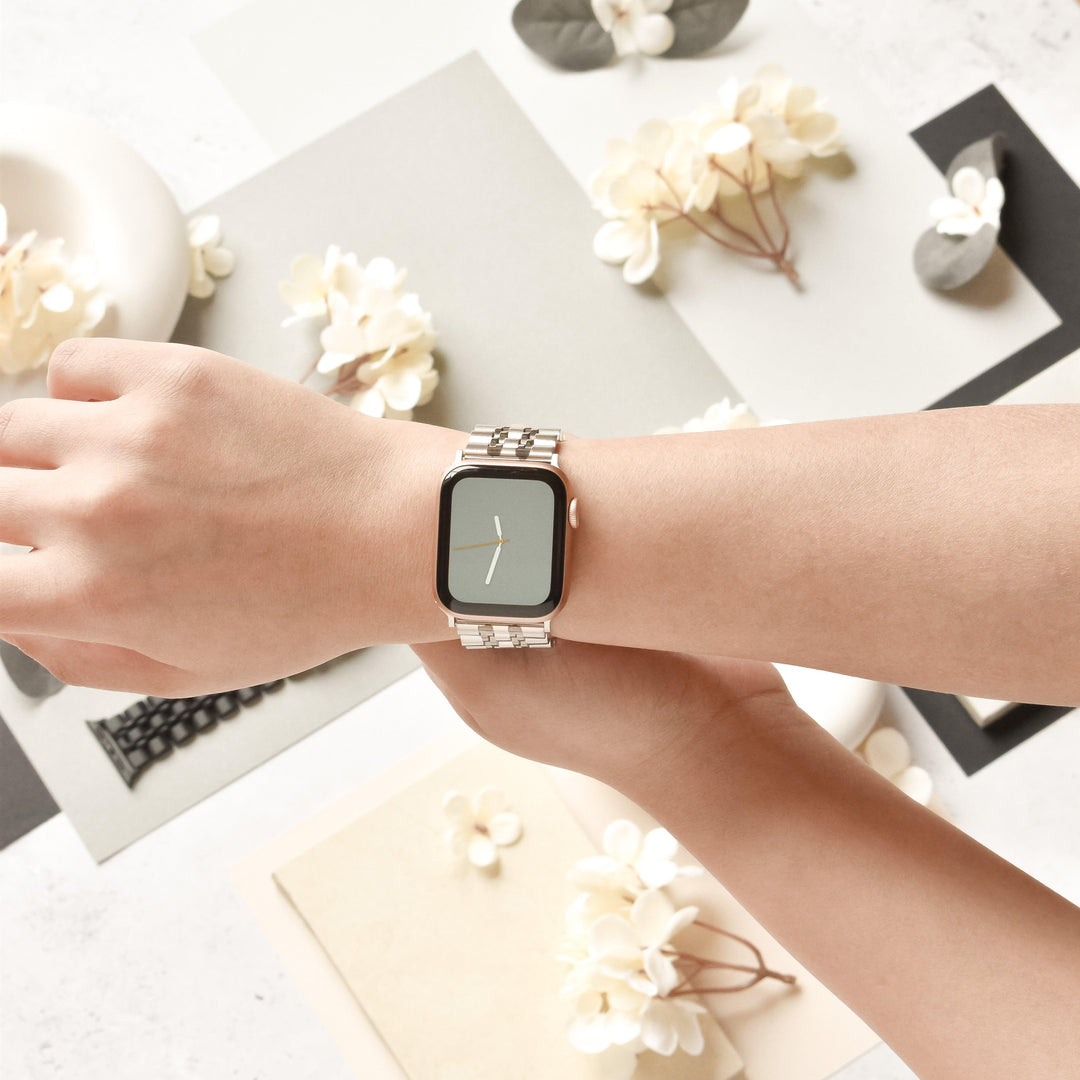 A person’s wrist is adorned with a sleek, metallic smart watch featuring a blank screen and the Coconut Lane Stainless Steel Apple Watch Strap - Black & Silver. The background is decorated with paper flowers, tasteful elements, and fabric swatches in neutral tones. The overall scene exudes a soft and stylish aesthetic, highlighting the elegance of the stainless steel strap as the focal point.