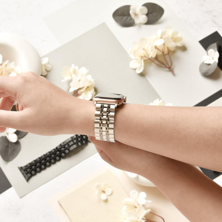 A person wearing a minimalist Coconut Lane Stainless Steel Apple Watch Strap in Black & Silver on their wrist, adjusting its strap with their other hand. The background features a flat lay of neutral-toned papers, white ceramic accessories, and scattered white flowers, creating an elegant and serene aesthetic.