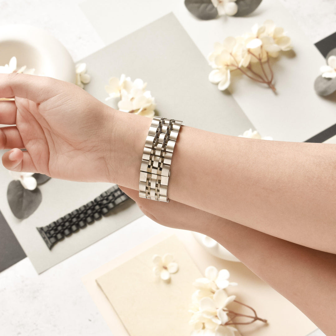 A person is trying on a Coconut Lane Stainless Steel Apple Watch Strap - Black & Silver on their wrist. The setup features a variety of watch bands, flowers, and small decorative items on a light-colored surface. The scene is bright and elegantly arranged.