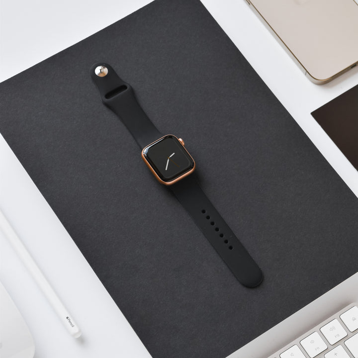 A stylish black smartwatch with the NAKD Apple Watch Strap - Midnight Black from Coconut Lane, featuring a minimalist watch face with a single white line, sits on a black rectangular surface. Nearby are parts of a white keyboard, a white pen, and a light-colored laptop, all neatly arranged to create a modern and organized workspace.