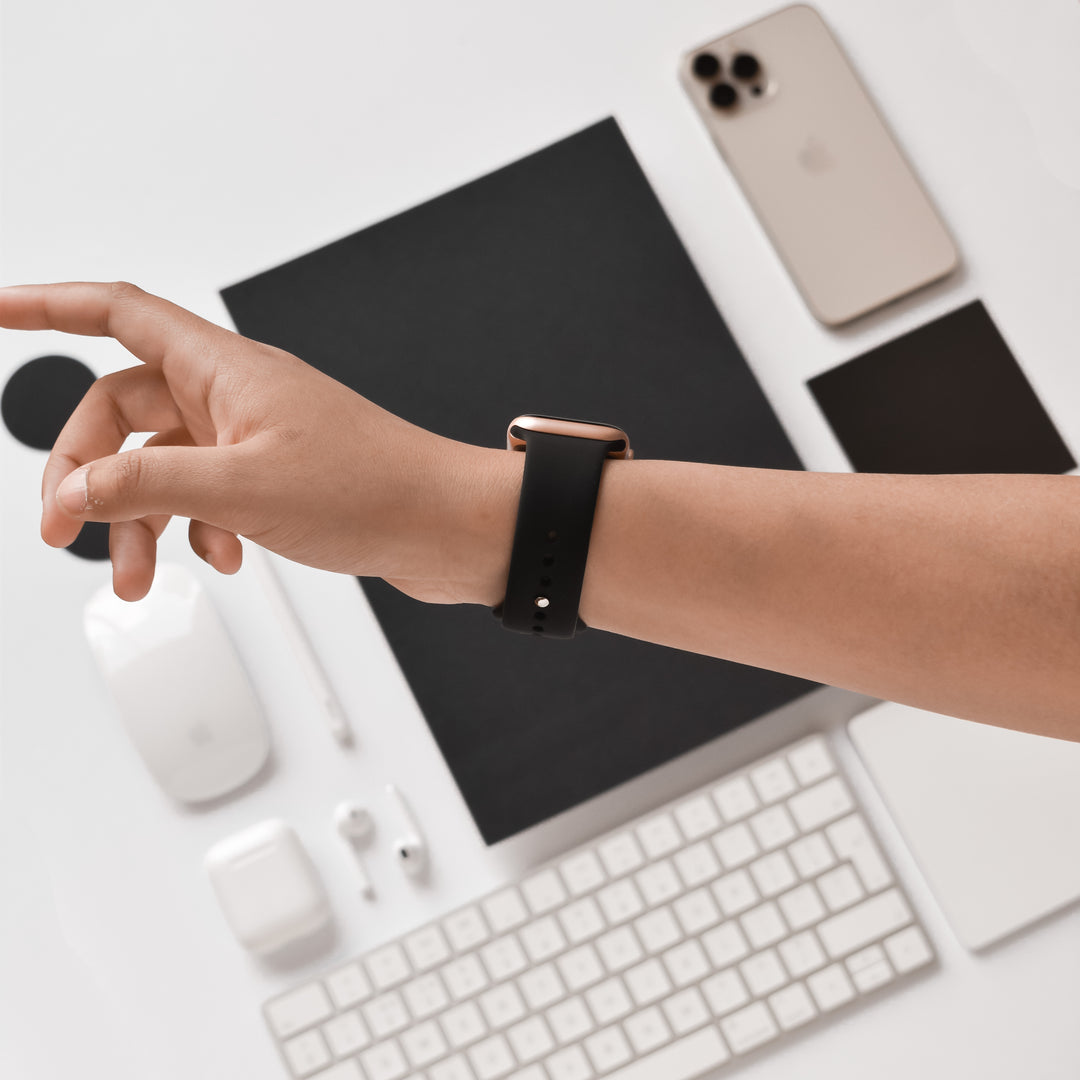 A person's hand, wearing the NAKD Apple Watch Strap - Midnight Black by Coconut Lane, is reached out over a white desk. On the desk are a keyboard, a wireless mouse, earbuds with their case, smartphone, small notepad, and various tech accessories from the NAKD collection in an organized layout.