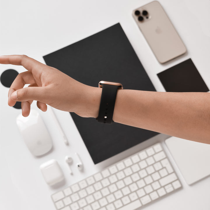 A person's hand, wearing the NAKD Apple Watch Strap - Midnight Black by Coconut Lane, is reached out over a white desk. On the desk are a keyboard, a wireless mouse, earbuds with their case, smartphone, small notepad, and various tech accessories from the NAKD collection in an organized layout.