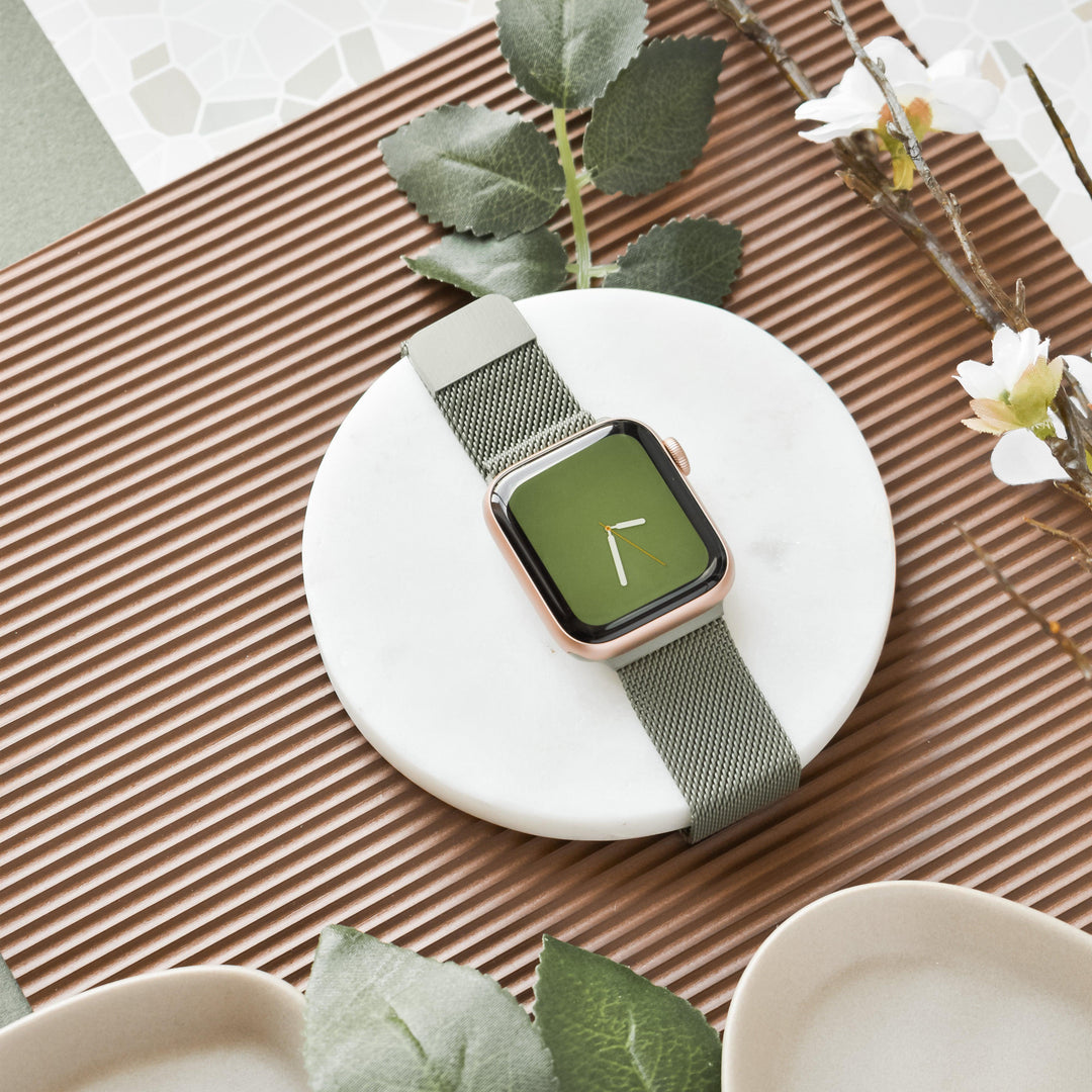 A sleek wristwatch with a green watch face and matching khaki mesh band is elegantly displayed on a white marble coaster. Surrounding the watch are decorative elements, including textured brown surfaces, leafy greenery, and white flowers. The Magnetic Chain Apple Watch Strap - Khaki by Coconut Lane adds both style and functionality.