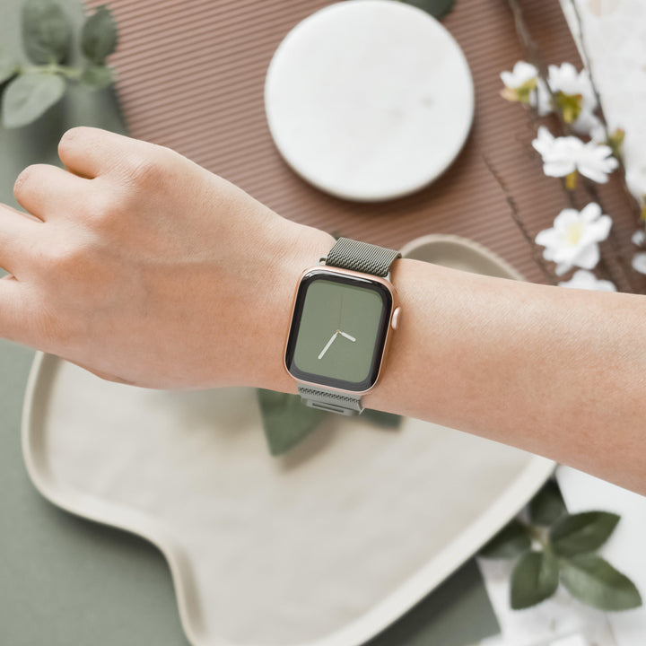 A person wearing a square-faced smartwatch with Coconut Lane's Magnetic Chain Apple Watch Strap in Khaki on their wrist. The smartwatch face features a minimalist design with a green background and white hour and minute hands. Their wrist is elegantly positioned over a decorative backdrop adorned with flowers and leaves.