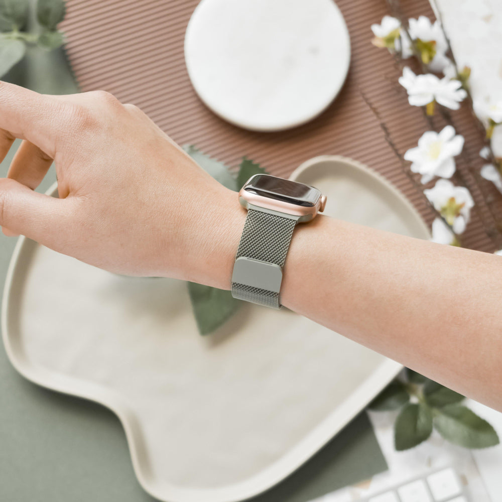 A hand wearing the Magnetic Chain Apple Watch Strap - Khaki from Coconut Lane rests on a neutral-colored, curved edge table. The table is adorned with white flowers, green leaves, and a round white and brown coaster. A keyboard is partially visible in the background. This stylish strap fits all Apple Watch models.