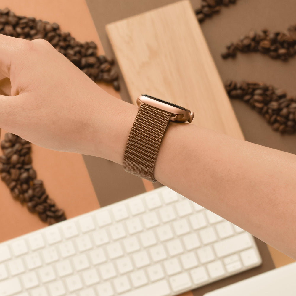 A close-up of a person's wrist wearing Coconut Lane's Magnetic Chain Apple Watch Strap in Coffee. The background features a mix of coffee beans arranged in patterns, a wooden board, and a white keyboard. The image seamlessly combines elements of tech, nature, and aesthetics.