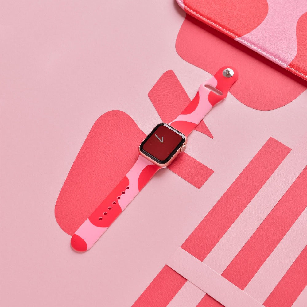 A smartwatch with an Abstract Pink & Red Apple Watch Strap is shown against a pink background adorned with various abstract shapes. Its screen features a minimalist red design with a white check-mark. A portion of a pink rectangular object and pink stripes are visible in the background, accentuating the Apple Watch strap by Coconut Lane.