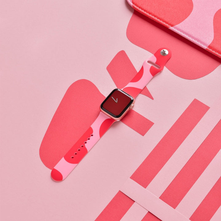 A smartwatch with an Abstract Pink & Red Apple Watch Strap is shown against a pink background adorned with various abstract shapes. Its screen features a minimalist red design with a white check-mark. A portion of a pink rectangular object and pink stripes are visible in the background, accentuating the Apple Watch strap by Coconut Lane.