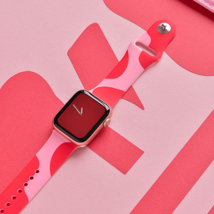 An Abstract Pink & Red Apple Watch Strap by Coconut Lane adorns a smartwatch resting on a pink surface. The watch face showcases a sleek red screen with white clock hands. Abstract pink shapes in the background enhance the cohesive aesthetic of the image.