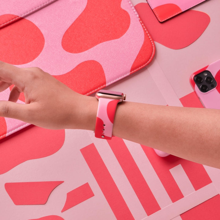 A person's forearm adorned with a Coconut Lane Abstract Pink & Red Apple Watch Strap is extended over a collection of items, including cases and covers that all feature matching designs. A smartphone in a pink case is also visible.
