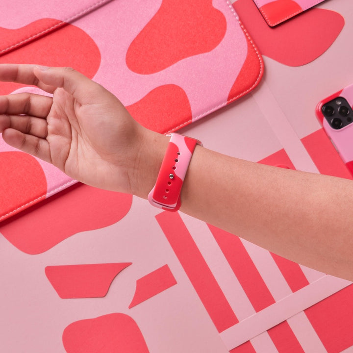 A person wearing an Abstract Pink & Red Apple Watch Strap by Coconut Lane rests their arm on a pink and red Coconut Lane patterned background. The scene includes items like a laptop sleeve, a smartphone, and a notebook, all in matching pink and red tones.