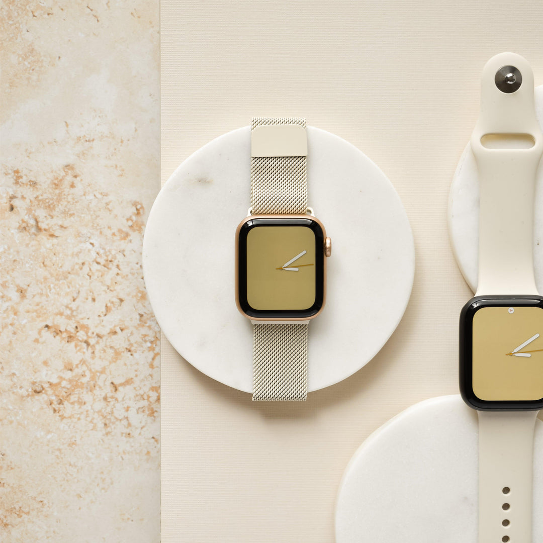 A Coconut Lane Apple Watch with an almond white magnetic chain strap and a golden face is elegantly displayed on a round marble slab, epitomizing metallic minimalism. Nearby, another Apple Watch with a light-colored silicone band and golden face rests on a flat, round surface. The background features a neutral, beige texture.
