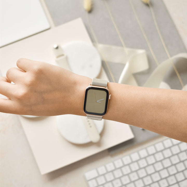 A person wearing a silver smart watch with a blank screen on their wrist, featuring the sleek Magnetic Chain Apple Watch Strap - Almond White from Coconut Lane. Below the wrist, there is a white keyboard, a white ribbon, square coasters, and other minimalistic stationery items on a beige surface.