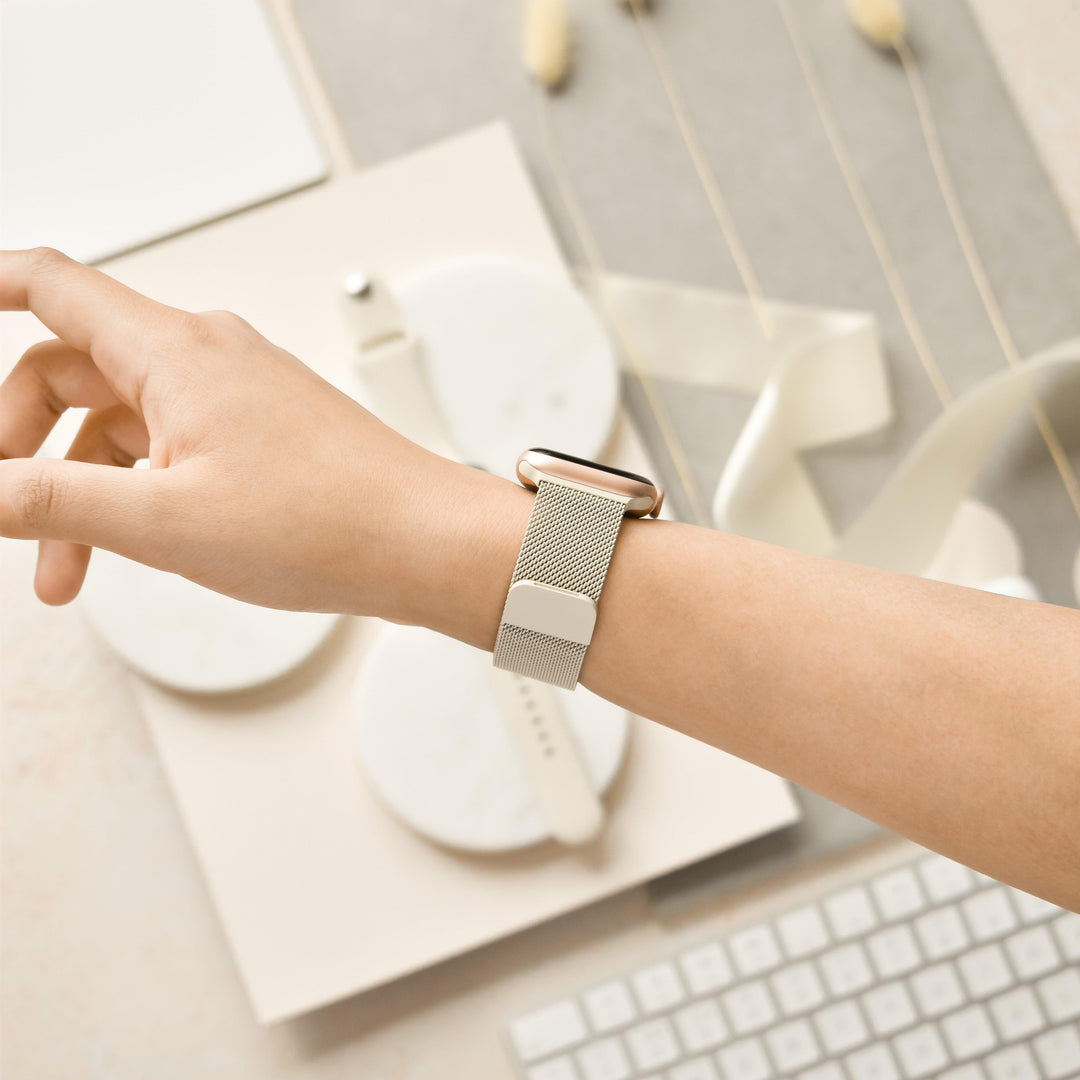 A close-up of a person's wrist wearing a smartwatch with the Coconut Lane Magnetic Chain Apple Watch Strap in Almond White. The background showcases a minimalistic workspace adorned with a keyboard, notebook, white circular objects, and a ribbon, all in soft, neutral tones.