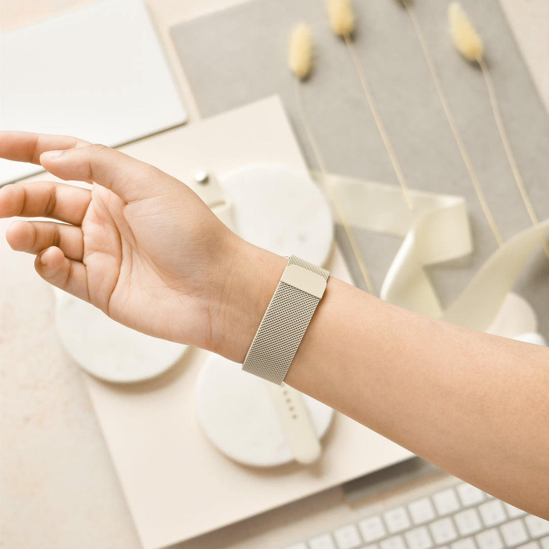 A close-up of a person's wrist adorned with the Magnetic Chain Apple Watch Strap - Almond White from Coconut Lane. The strap's sleek, metallic mesh design is complemented by a stainless steel adjustable clasp. In the background, a stylish, minimalistic setup features a keyboard, decorative twine, white round objects, and dried floral arrangements in soft, neutral tones.