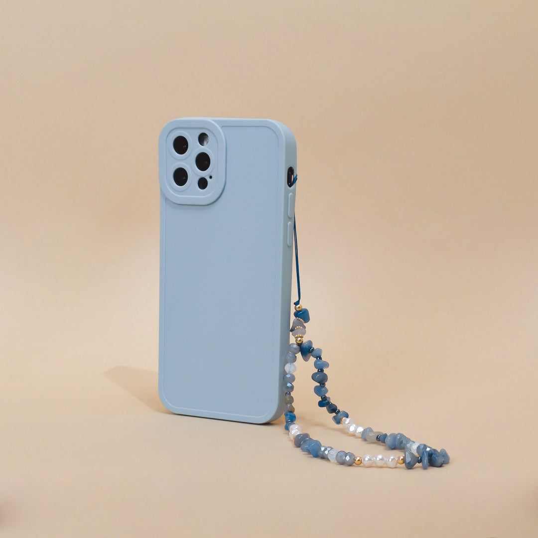 A light blue smartphone case with a camera cutout is displayed standing vertically against a beige background. Attached to the case is Coconut Lane's "Beaded Phone Strap - Ocean Eyes," made of blue beads and small pearls, adding an elegant touch to your phone accessories.