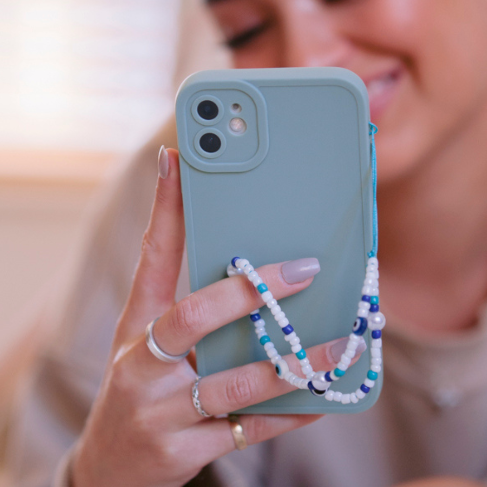 A person holding a smartphone with a teal case is adorned with the Beaded Phone Strap - Evil Eye by Coconut Lane, featuring beaded charms in various shades of blue and white. The person is smiling in the blurred background, wearing silver rings on their fingers.