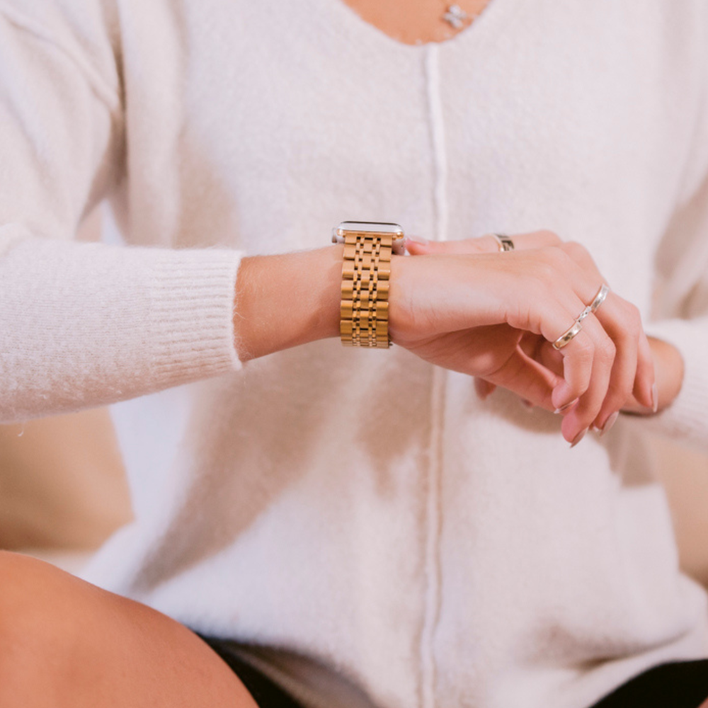 A person wearing a white sweater is looking at a Coconut Lane Stainless Steel Apple Watch Strap - Gold on their wrist. They have rings on their fingers and are holding their wrist with their other hand. The background is out of focus, highlighting the watch and hands.
