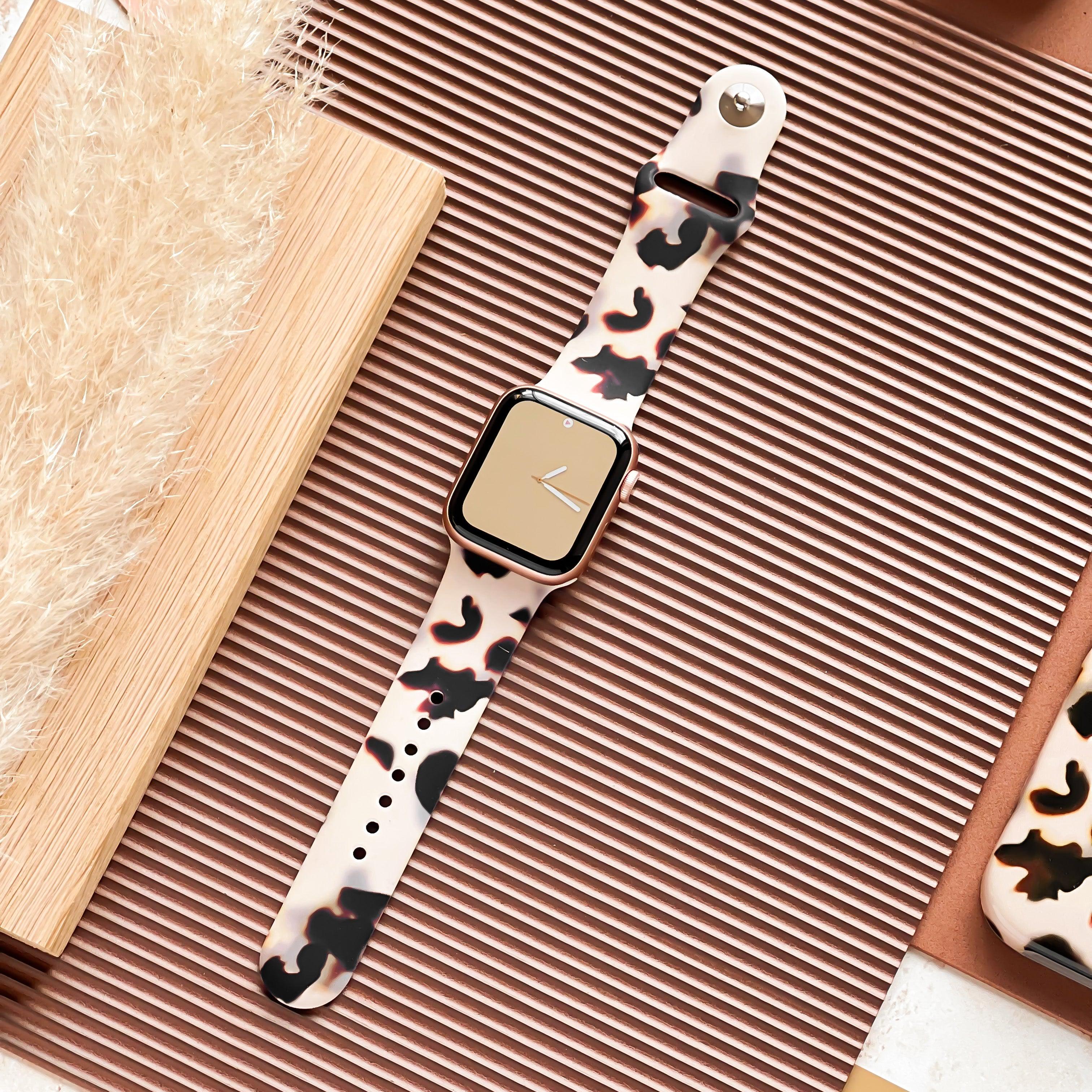 A smartwatch with a beige face and Coconut Lane's Milky Ivory Tort Apple Watch Strap featuring black and brown cow print spots lies on a textured, light brown surface next to a wooden board and a plant with beige feathery leaves. The background includes various brown tones and a partial view of a phone case.