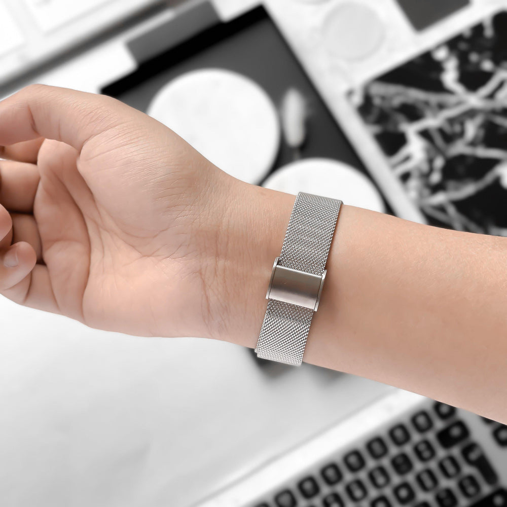 A close-up of a person's wrist adorned with the sleek Milanese Apple Watch Strap in Silver by Coconut Lane. The background showcases a blurred-out laptop keyboard and monochrome abstract designs, drawing attention to the watch strap's minimalist and modern design, which is compatible with various Apple Watch models.