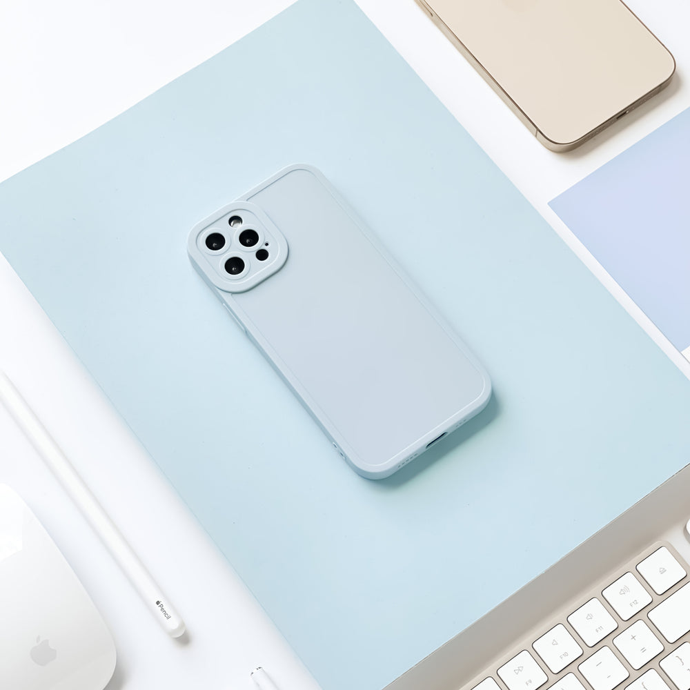 A light blue smartphone with a Coconut Lane NAKD Phone Case in baby blue lies on a blue paper background. Surrounding it are parts of a white computer keyboard, a white stylus, a gold smartphone, and other blue and white office supplies, creating a clean and minimalistic workspace setting.